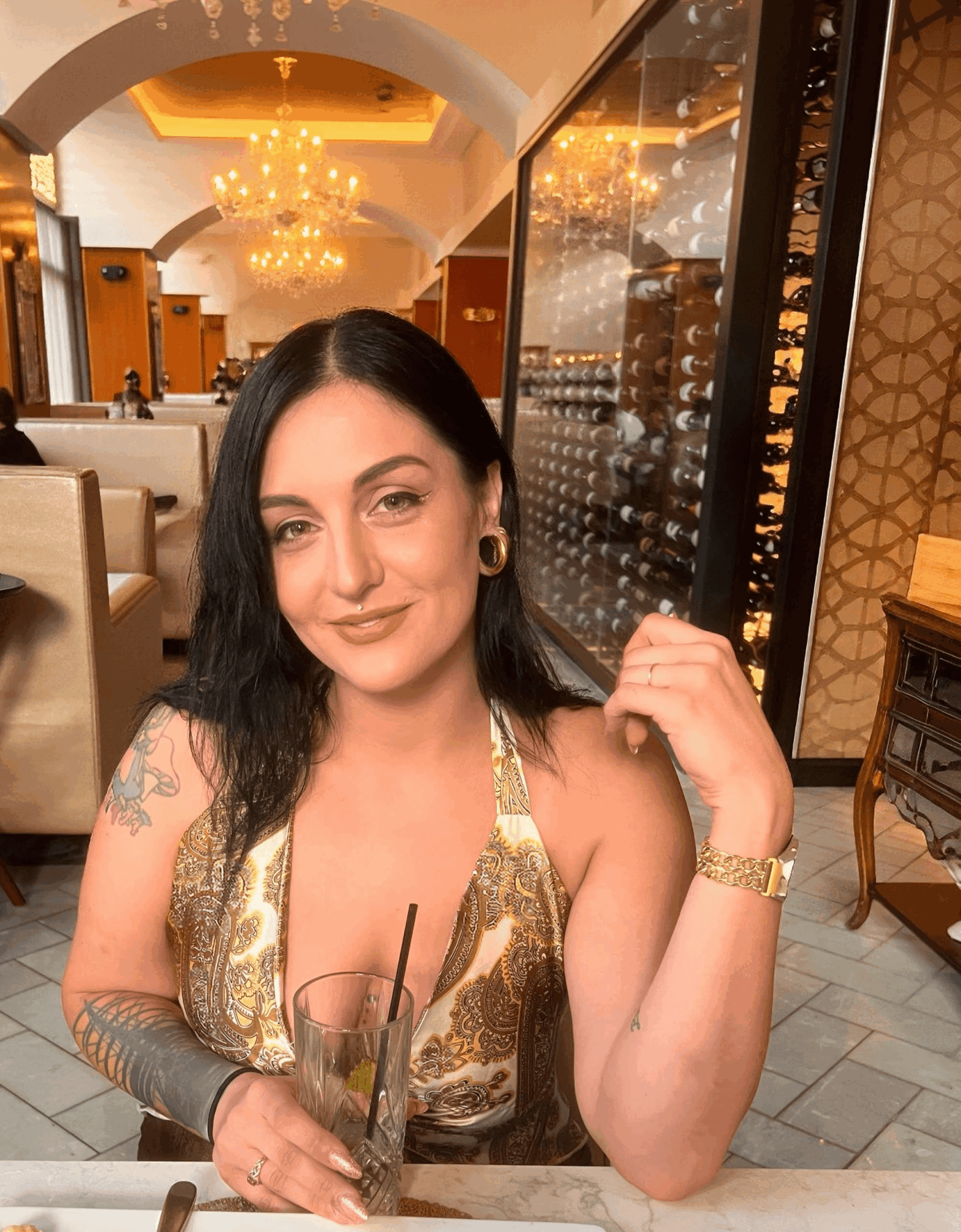 Woman with dark hair, gold dress, and jewelry sitting in a restaurant with chandeliers and glass display case behind her.