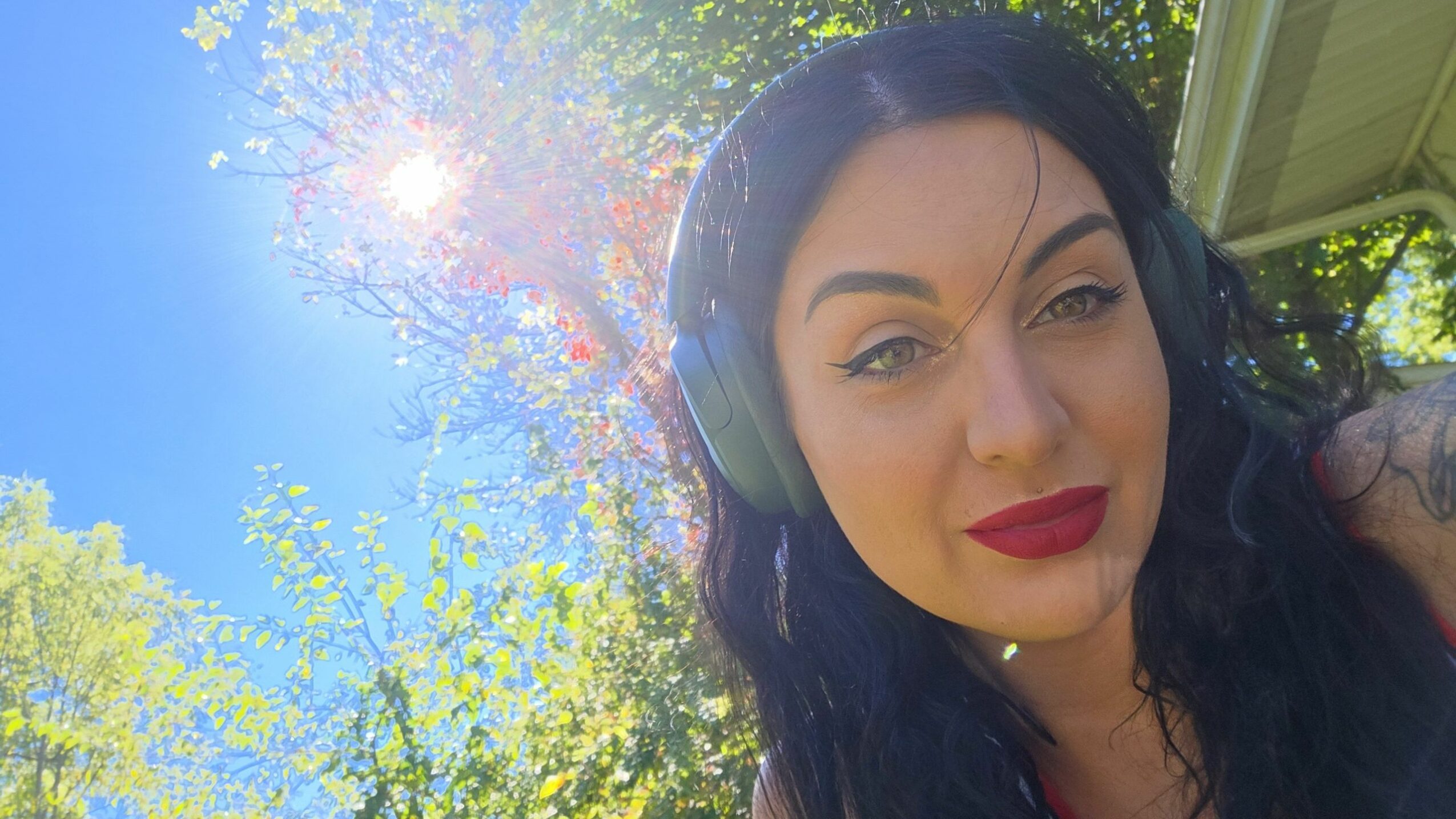 Woman with dark hair and red lipstick outdoors, blue sky and sunlight visible, trees in background.