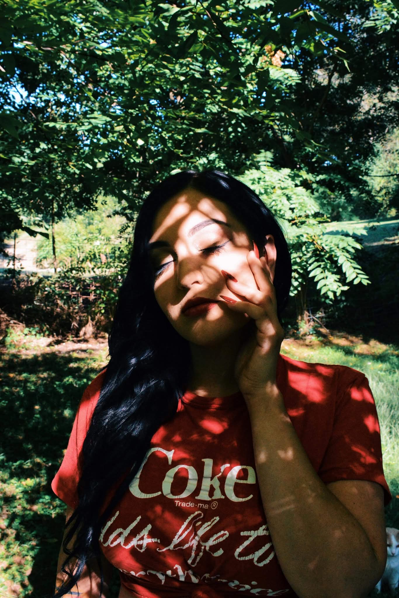 Young woman with long dark hair outdoors, sunlight casting shadows on her face, wearing a red shirt with white text, surrounded by trees.