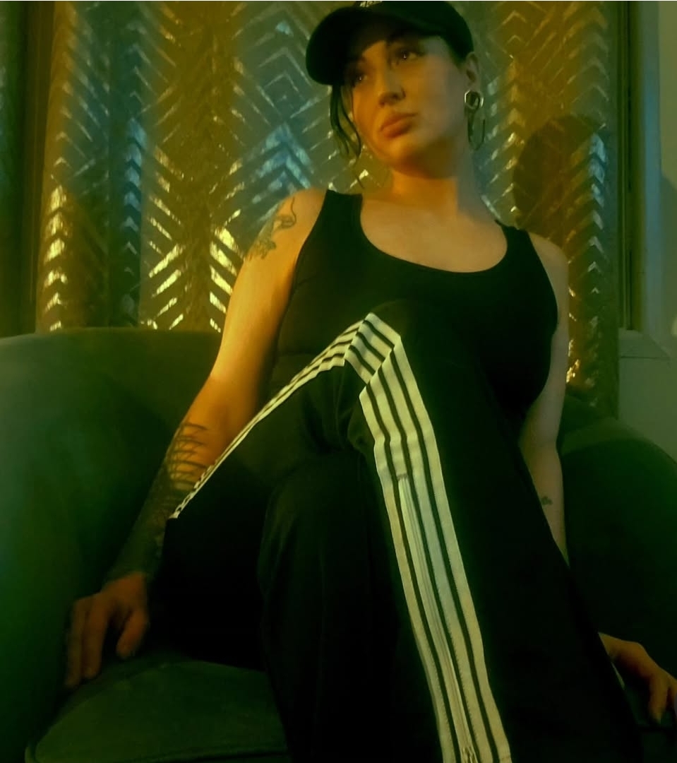 Woman sitting on a green couch, wearing a black tank top, striped pants, and a cap, with a patterned background.