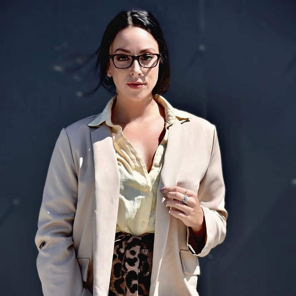 Woman with glasses and dark hair wearing a beige blazer and light-colored shirt, standing against a dark background.