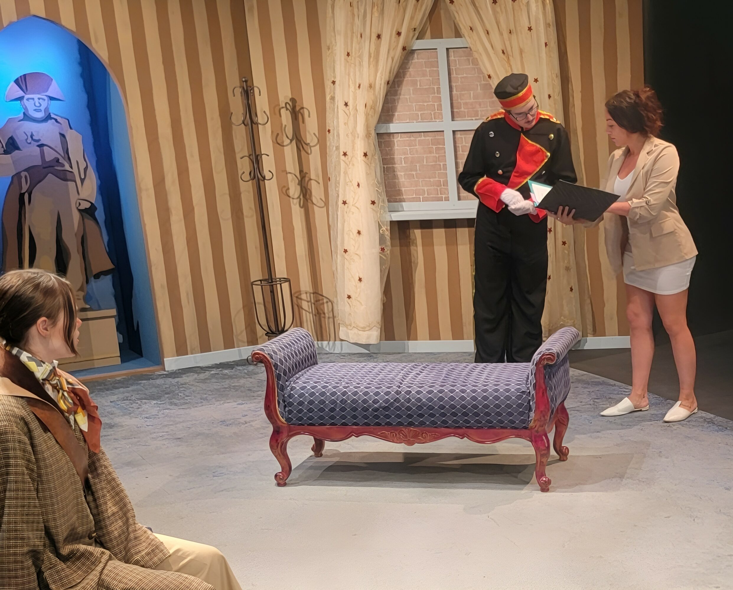 Stage scene with four people, a vintage sofa, and a window with curtains, set in a room with striped wallpaper.