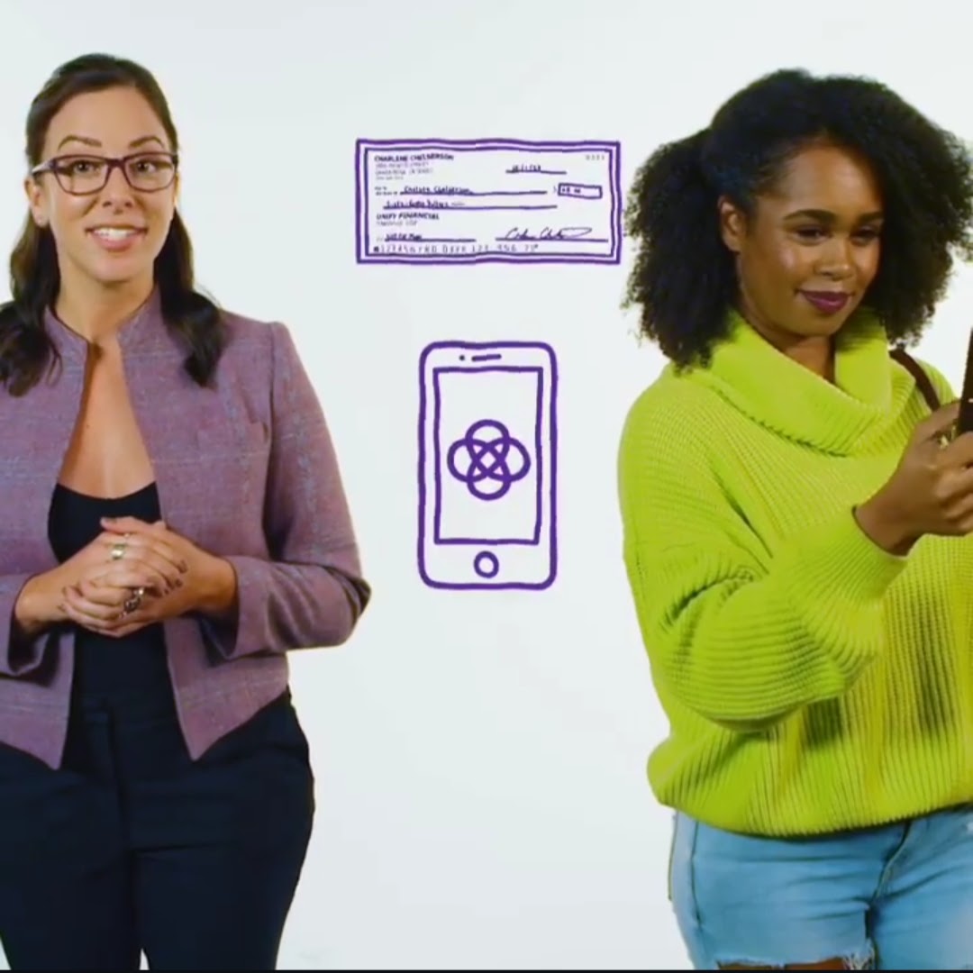 Two women standing, one smiling and the other looking at a phone, with sketches of a check and a mobile app on the wall behind them.