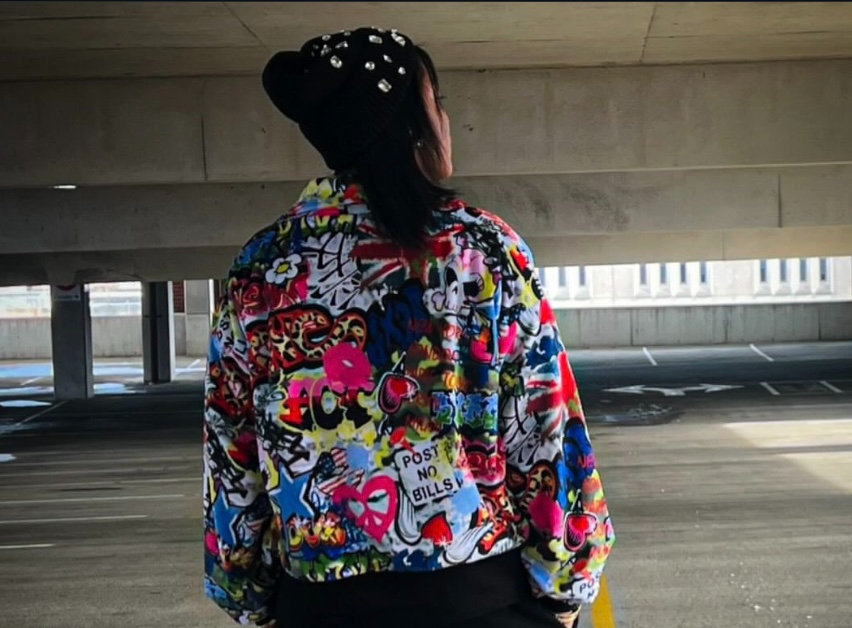 Person with dark hair in a colorful, patterned jacket, standing under a bridge, facing away from the camera.
