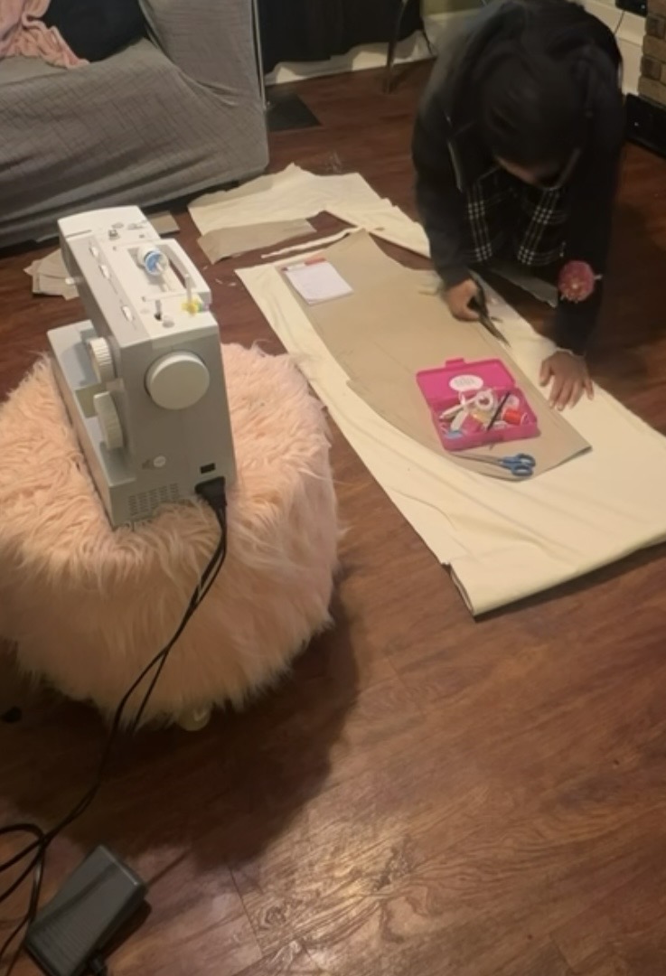 Person working on a sewing project on the floor with fabric, scissors, and a pink box, next to a sewing machine on a fluffy pink rug.