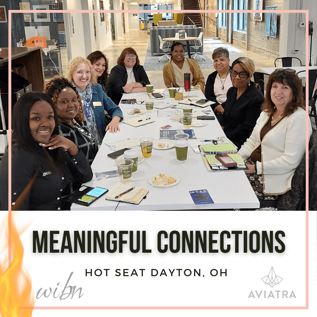 Group of women sitting around a table in a bright room, smiling, with notebooks and drinks, during a meeting or event.