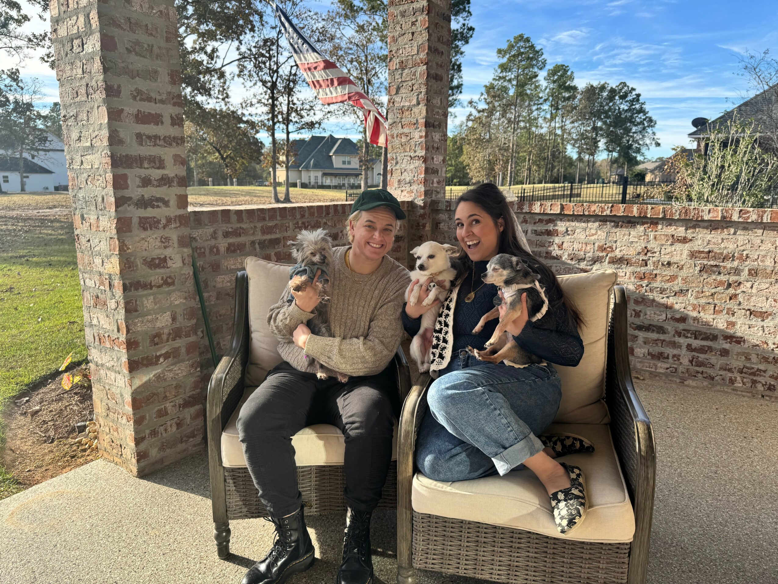 Two women sitting on outdoor bench with two dogs, brick wall and trees in background, sunny day.