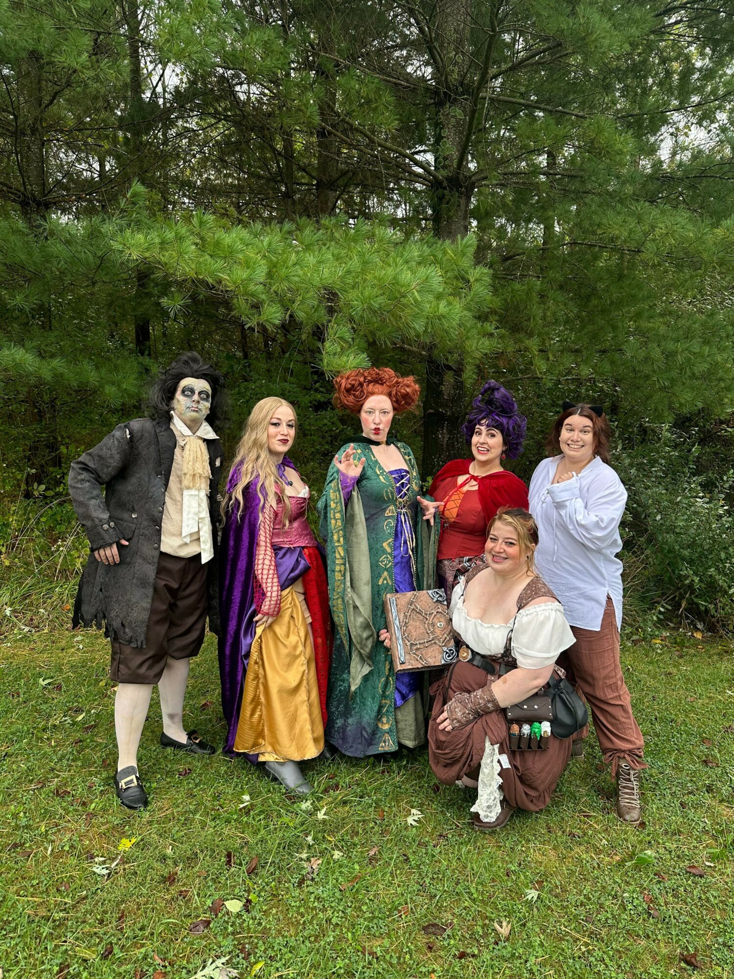 Group of six people standing outdoors on grass with trees in background, posing for photo.
