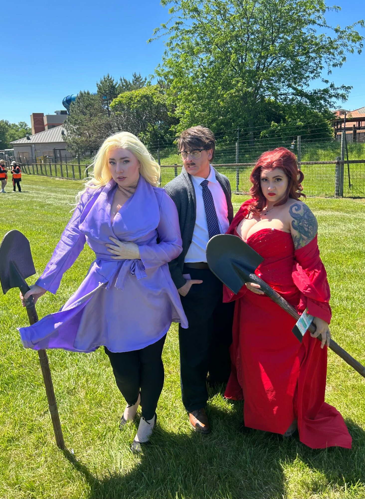 Three people in costumes standing on grass, holding shovels, with trees and a fence in the background.