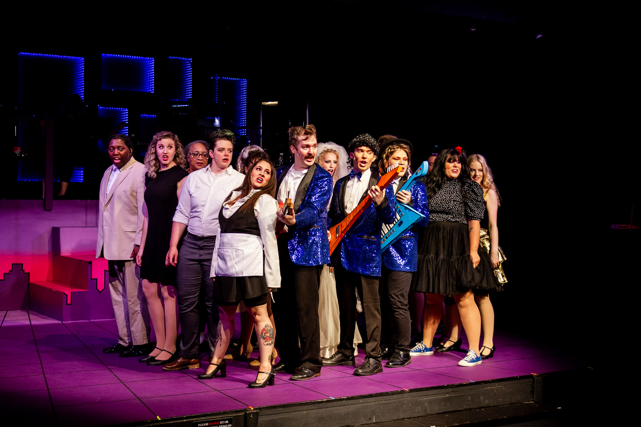 Group of people on stage, some holding musical instruments, with colorful lighting and set pieces in the background.