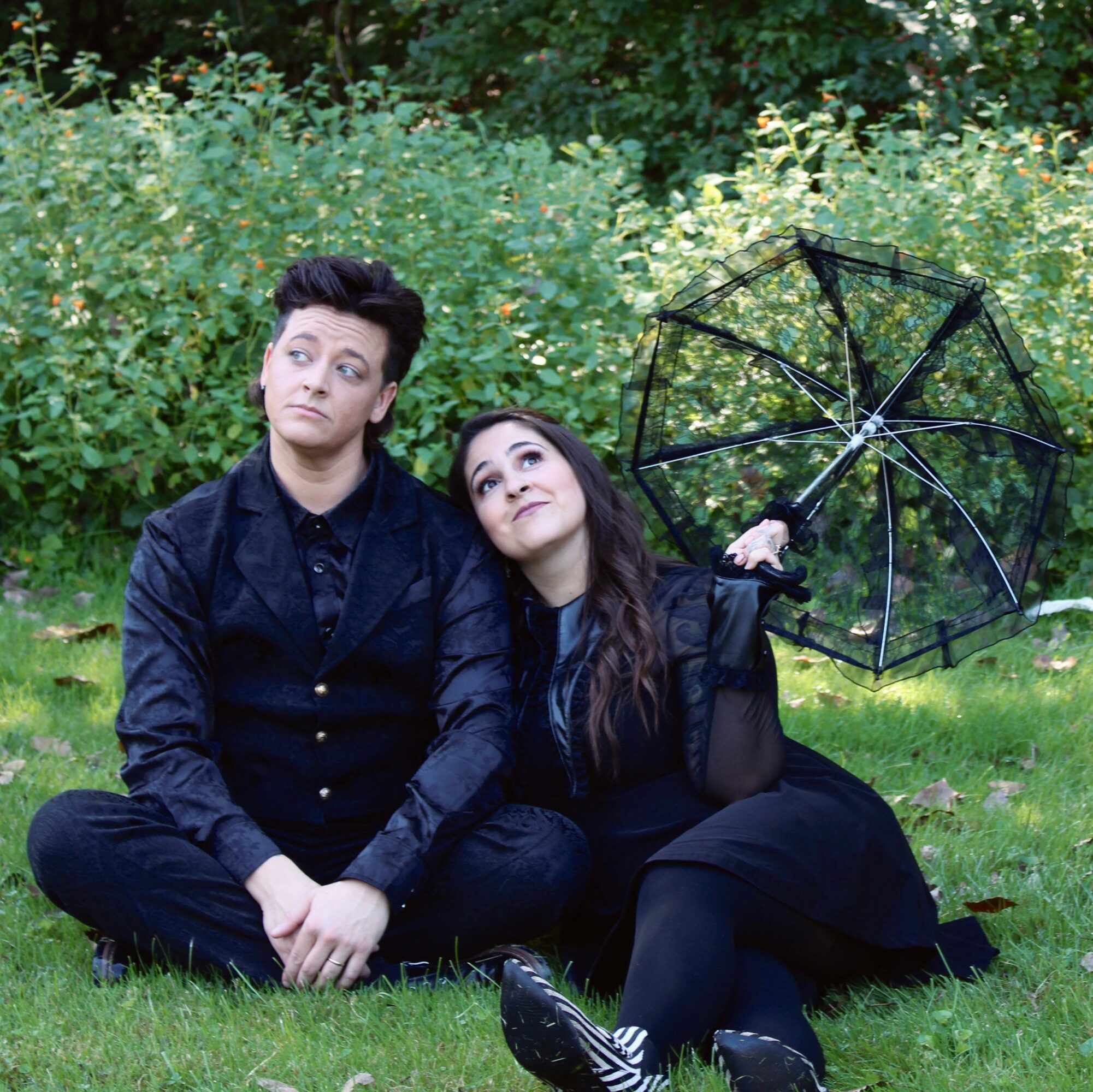 Two people sitting on grass with a black umbrella, surrounded by green bushes, looking upward thoughtfully.