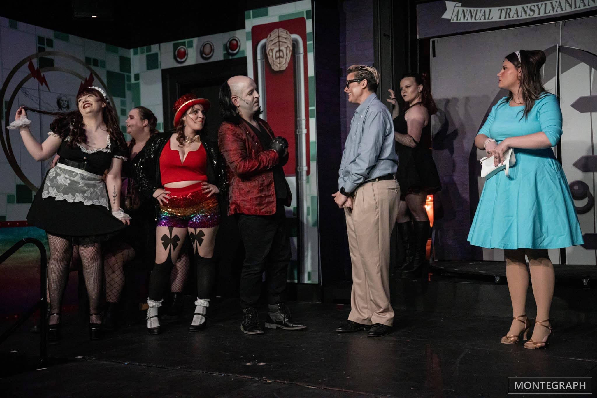 Group of people on stage, some in costumes, engaging in conversation or performance, with a decorated background and stage lighting.