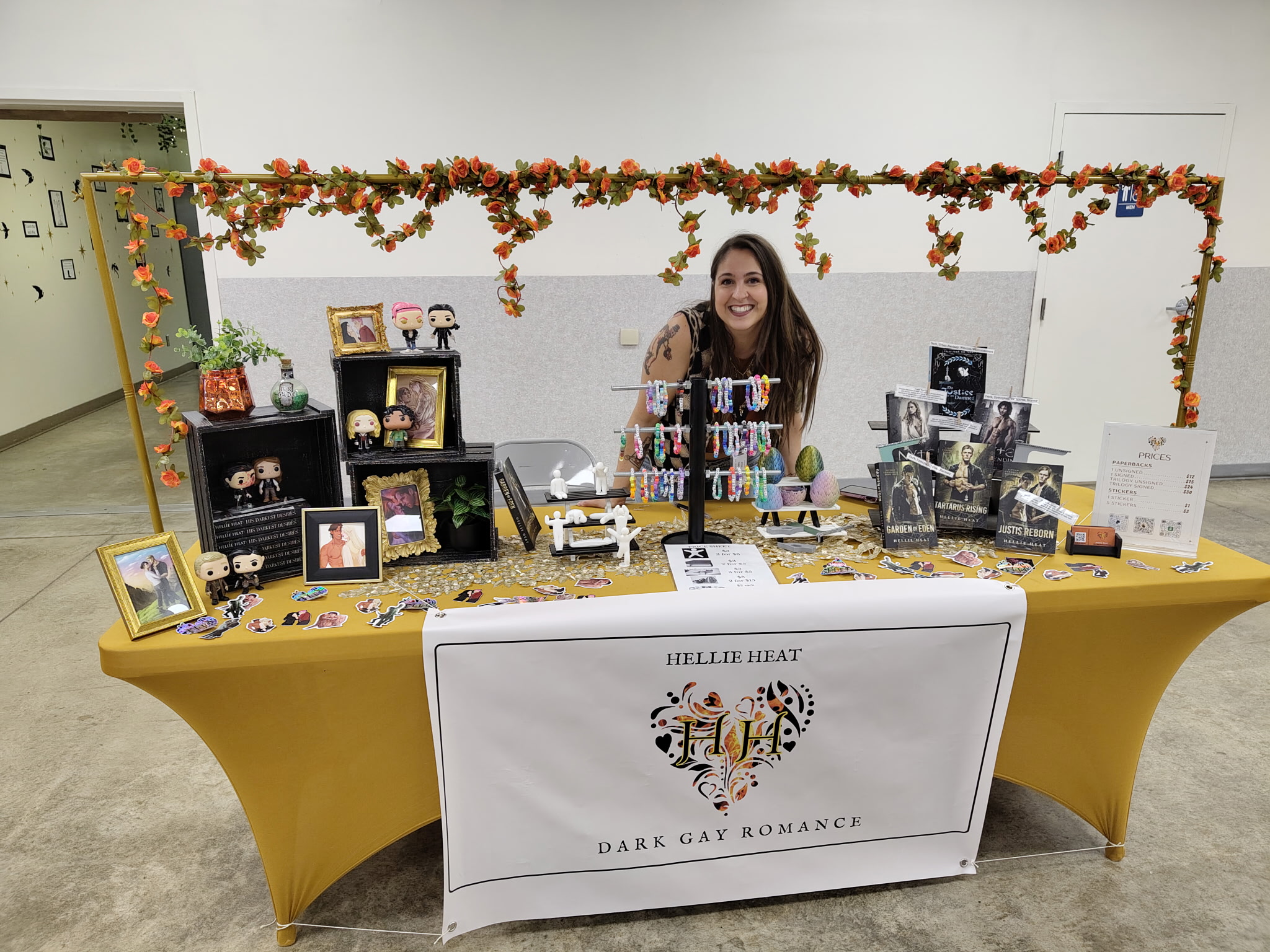 Woman standing behind a table with jewelry and accessories, decorated with a floral garland, in a display booth. Worded banner reads 'Hello Heat, Dark Gay Romance'.