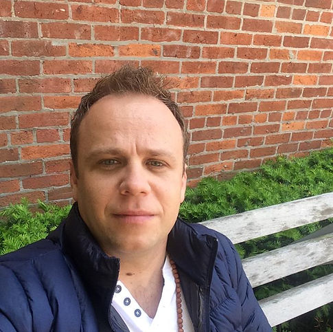 Man with short hair and light skin taking a selfie outdoors, brick wall and green grass in background.