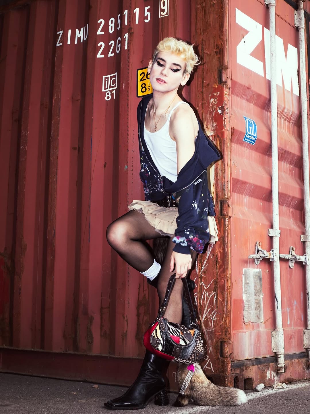 Young woman with blonde hair and makeup leaning against a red shipping container, holding a bag, with a dog at her feet.