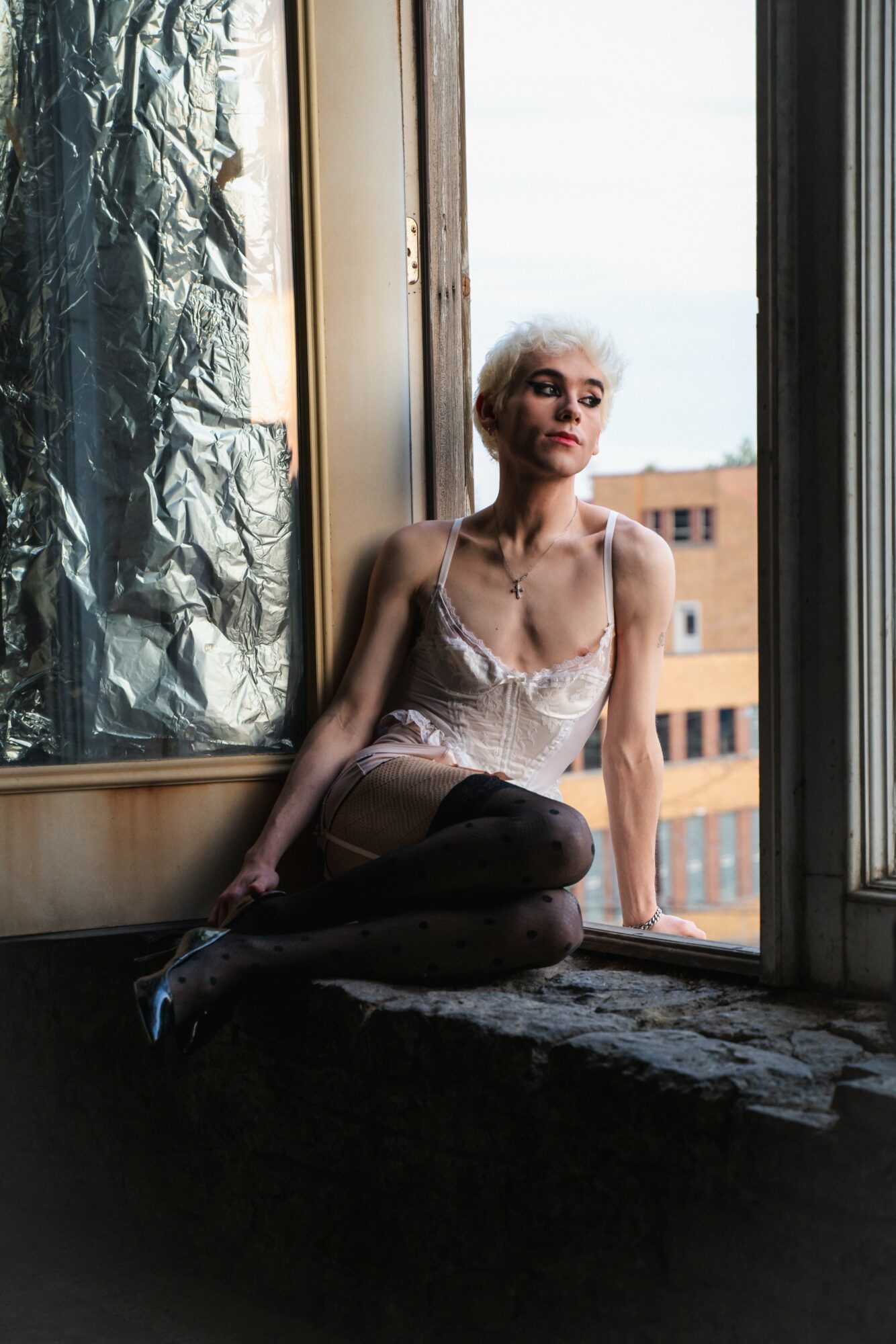 Young woman with short blonde hair sitting on windowsill, looking outside, wearing a light-colored top and dark pants.