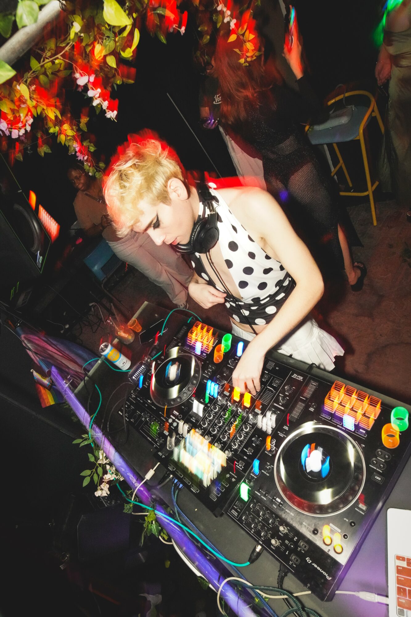 Young woman with short blonde hair wearing a polka dot top and headphones DJing at a colorful party.
