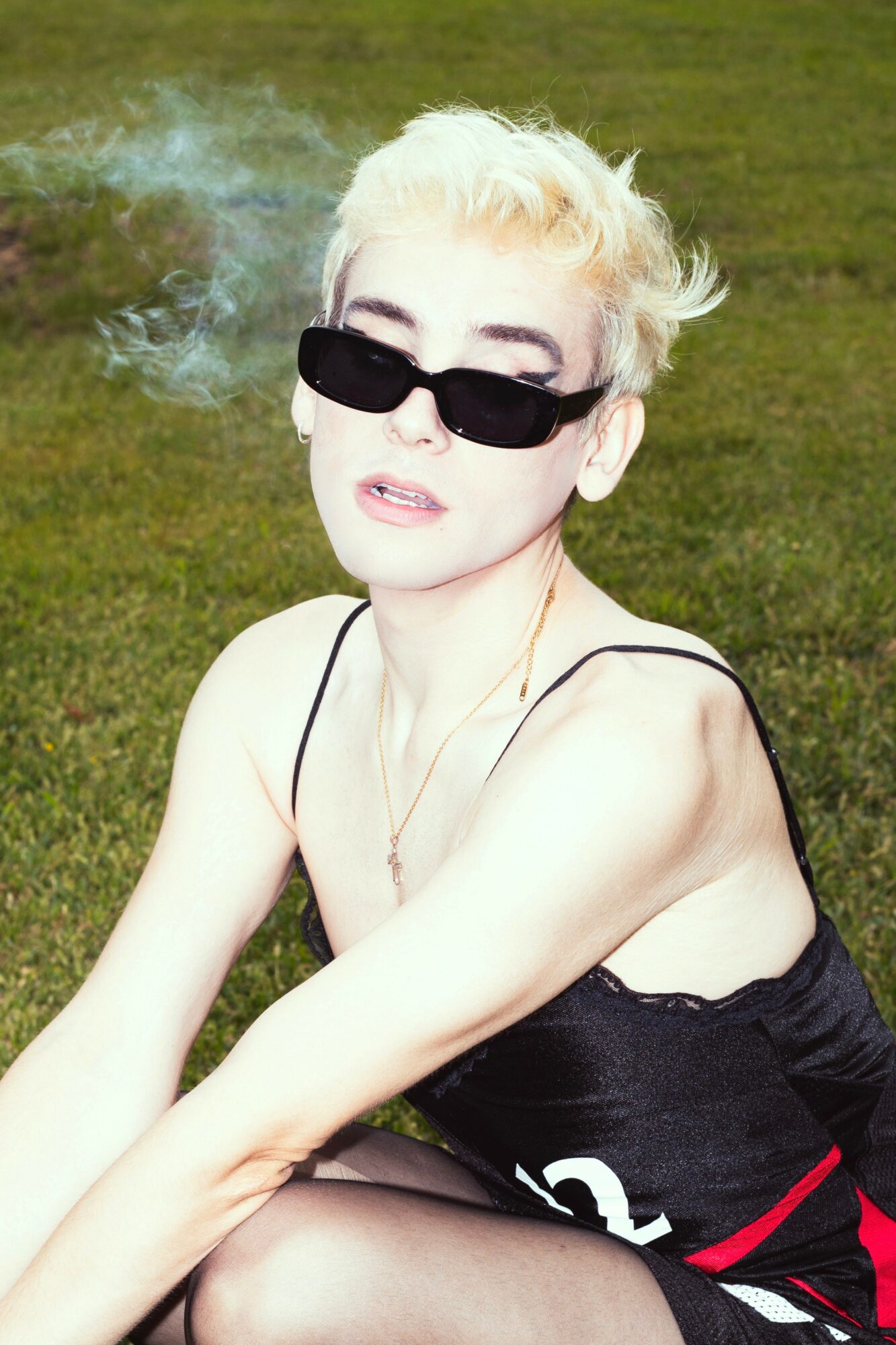 Person with short blonde hair wearing sunglasses, sitting outdoors on grass, wearing a black top, with smoke in the background.