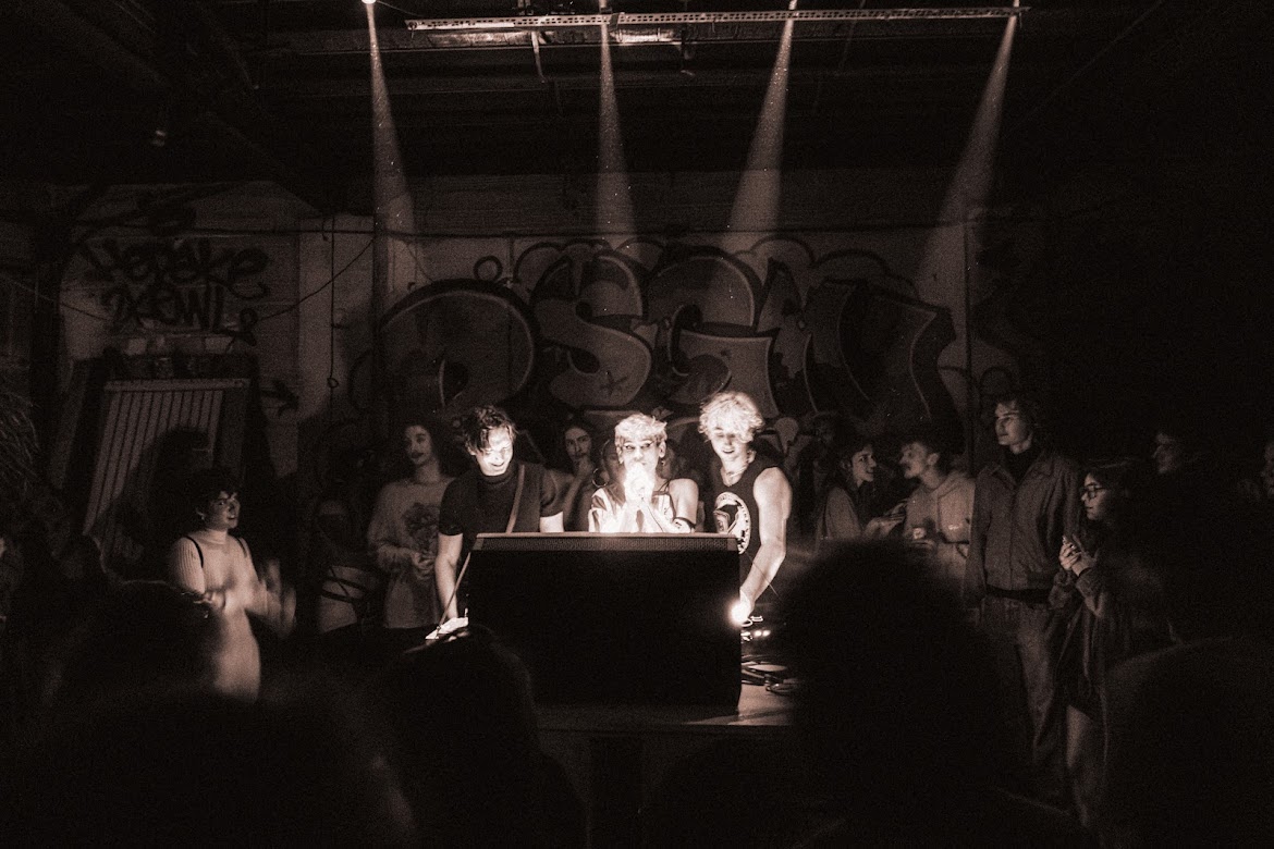 People gathered around a table in a dimly lit room with graffiti on the wall, some illuminated by overhead lights.