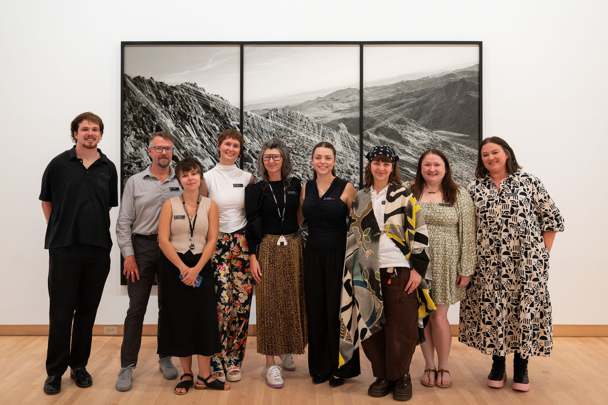 Group of nine people standing in front of a three-panel black and white landscape photograph, smiling, in a gallery.