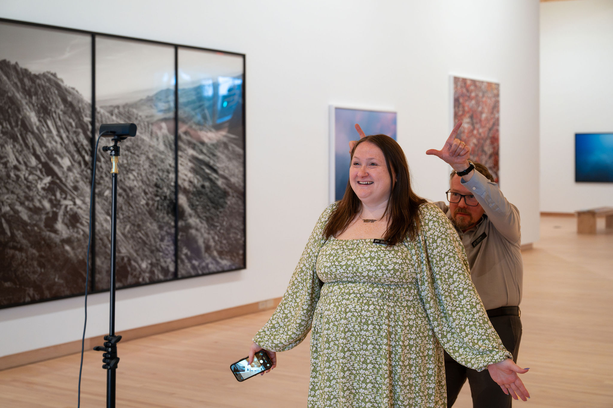 Woman with long hair smiling, holding a phone, standing in an art gallery with large landscape paintings on the wall.
