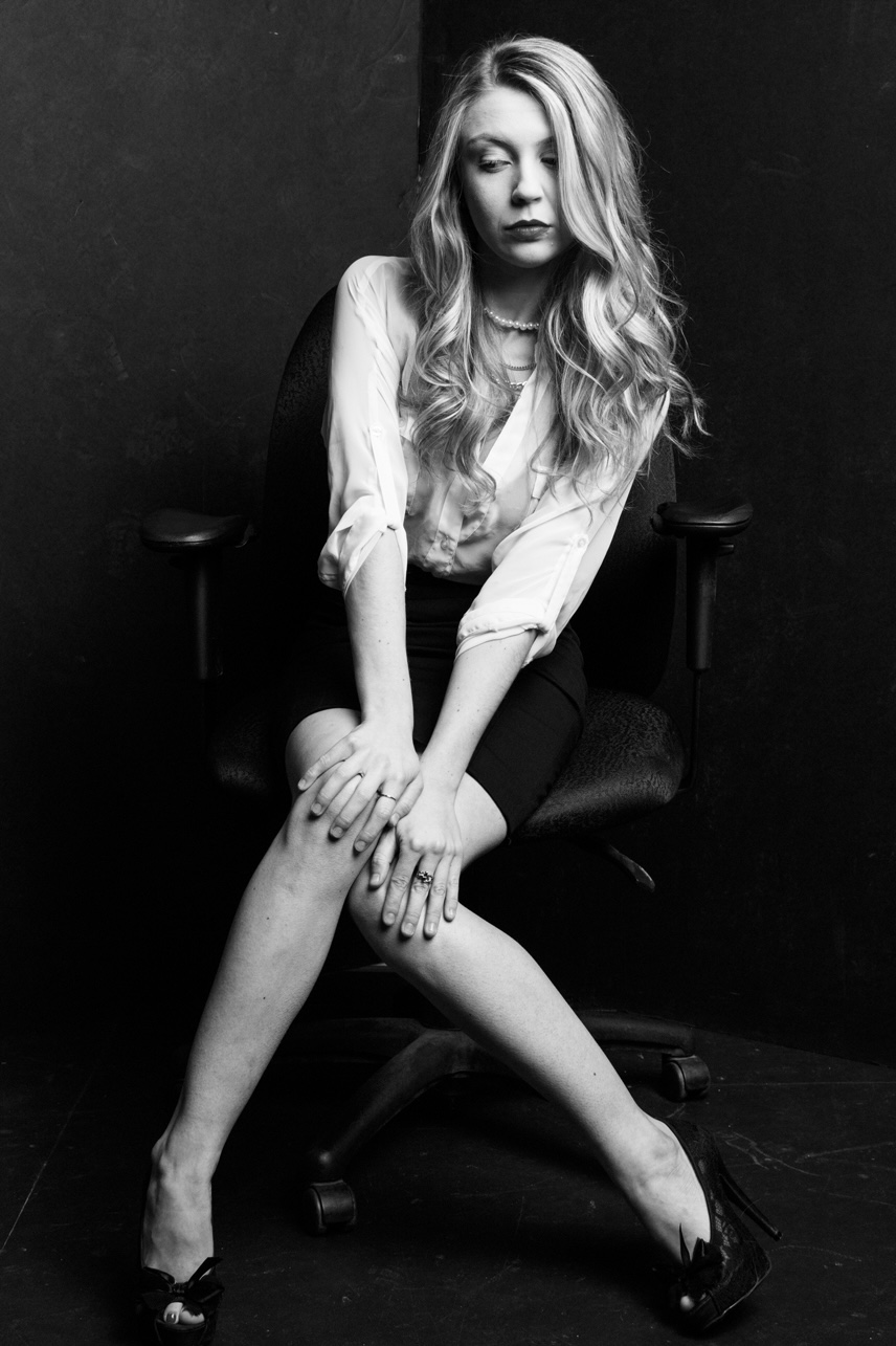 Woman with long hair sitting on a chair, hands on her knee, in black and white.