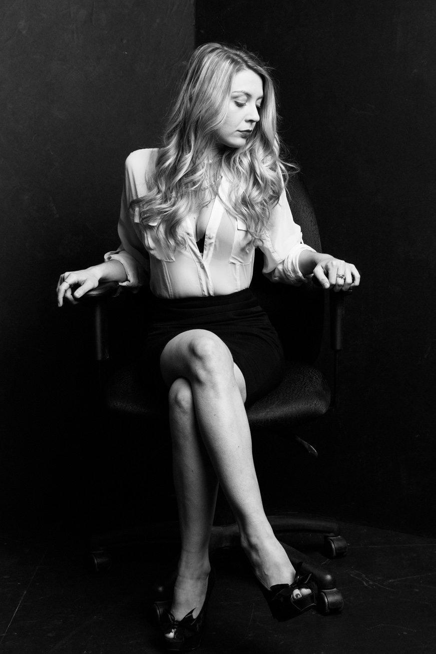 Woman with long hair sitting on a chair, looking down, in black and white, with a dark background.