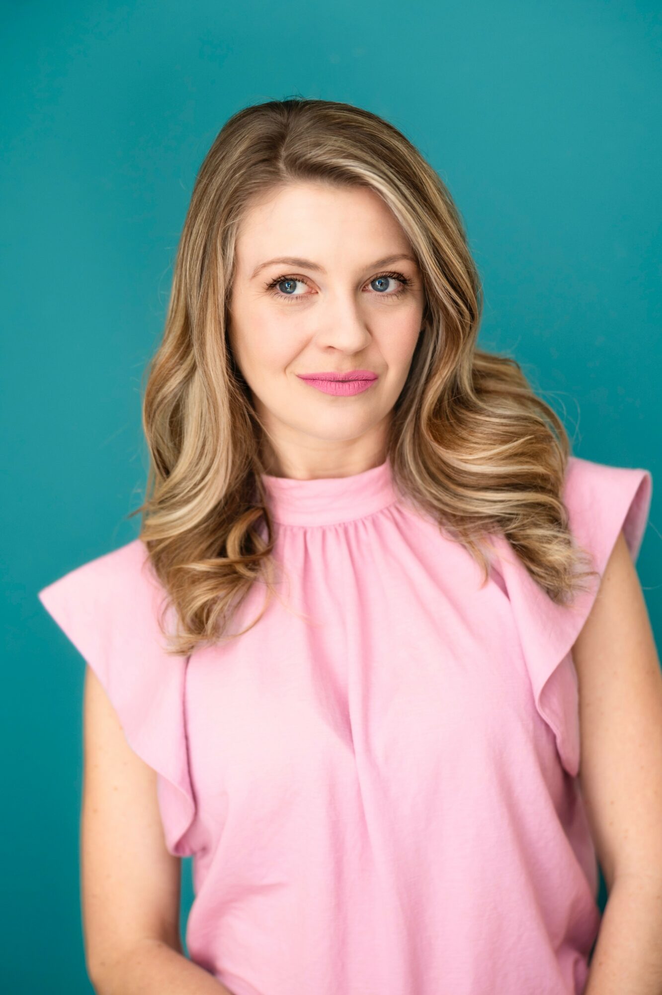Woman with wavy blonde hair wearing a pink top, smiling, against a teal background.