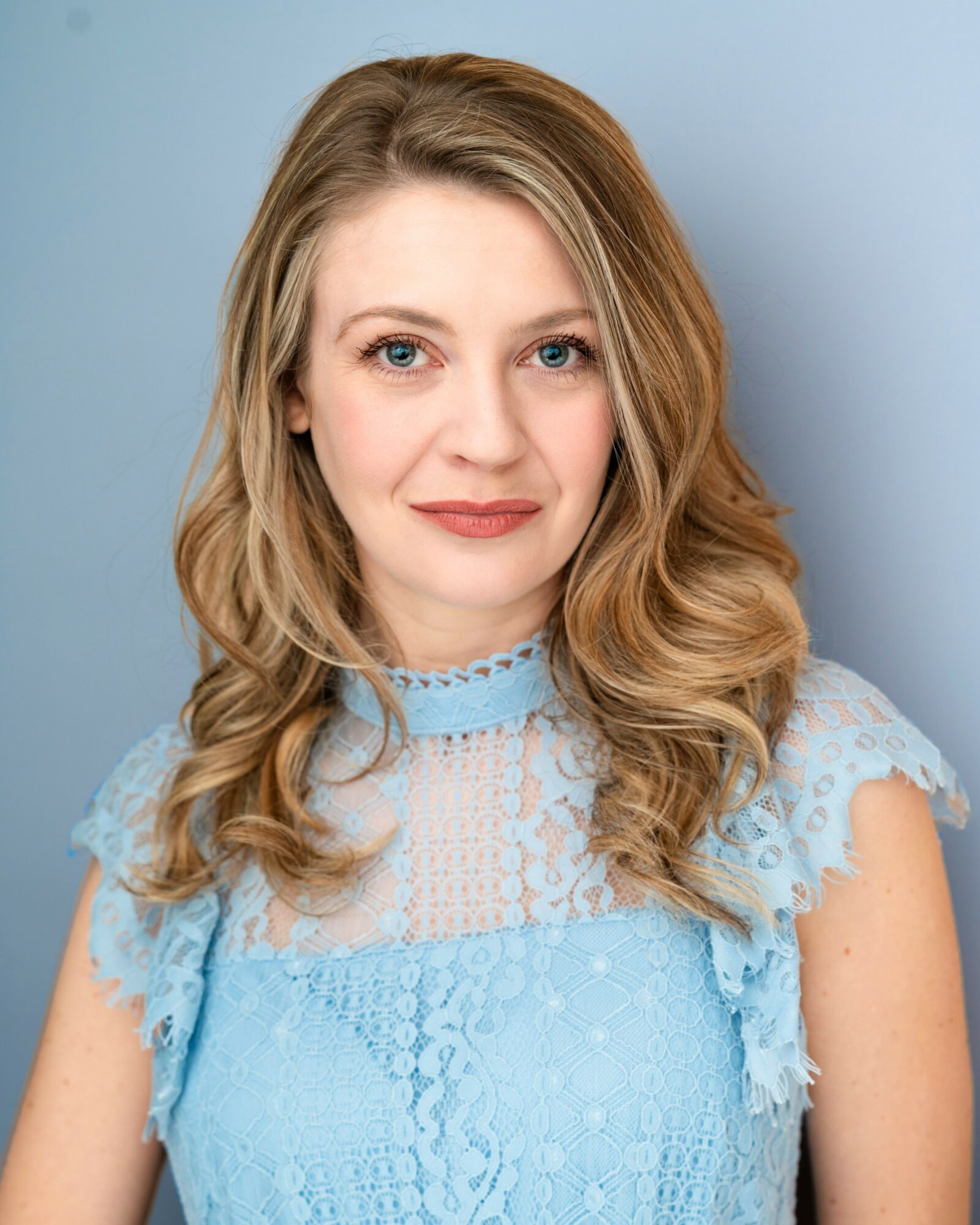 Woman with wavy blonde hair wearing a light blue lace top, smiling softly against a blue background.