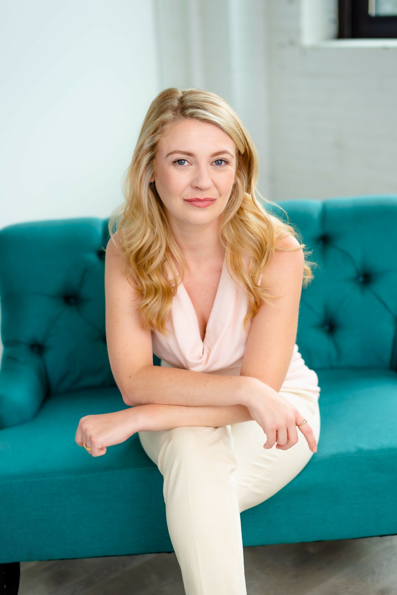 Young woman with blonde hair sitting on a teal sofa, wearing a light pink top and white pants, smiling softly.