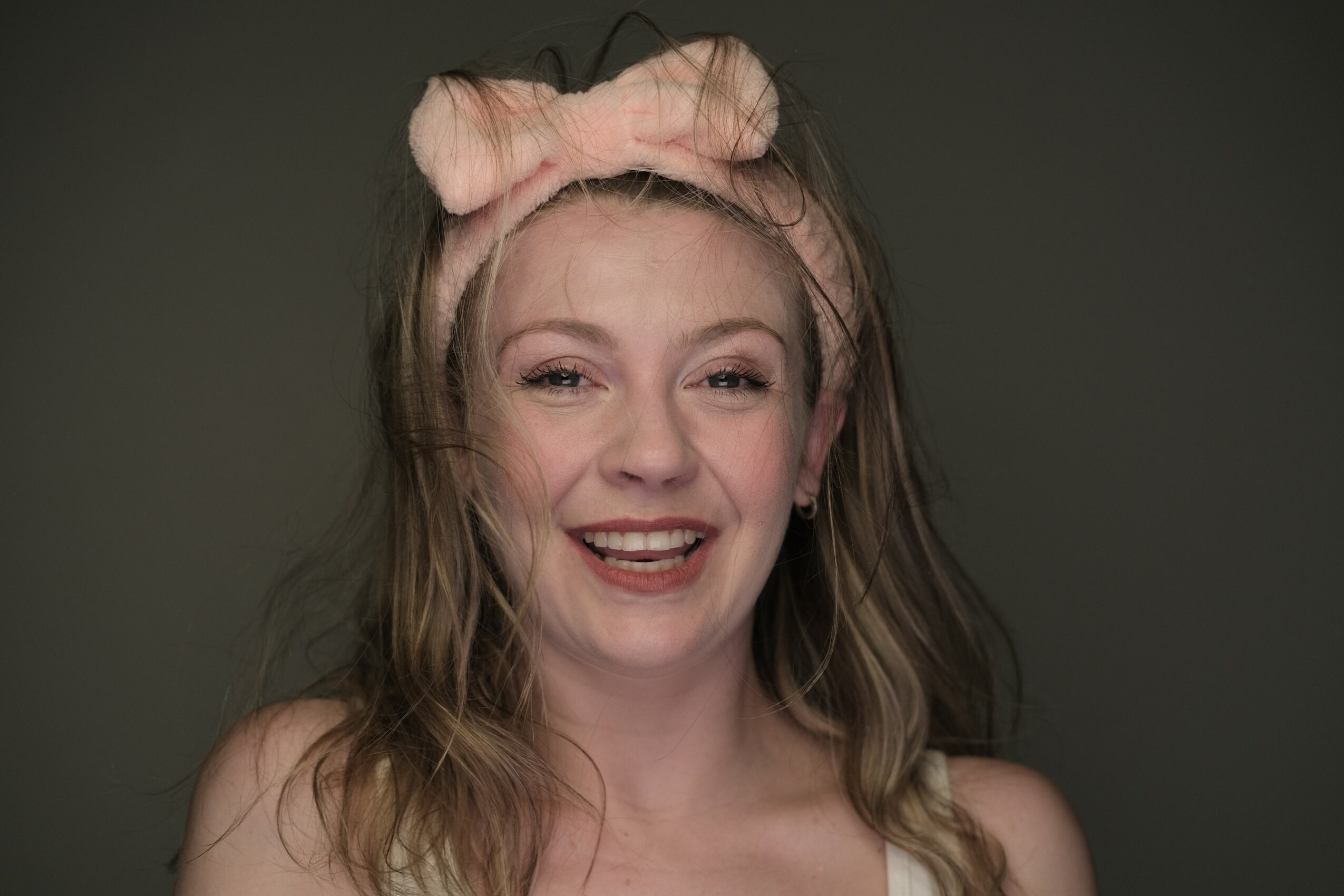 Young woman with wavy hair wearing a pink headband with a bow, smiling, against a dark background.