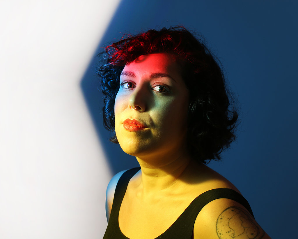 Portrait of a woman with curly hair, illuminated by colorful lighting, against a blue background.