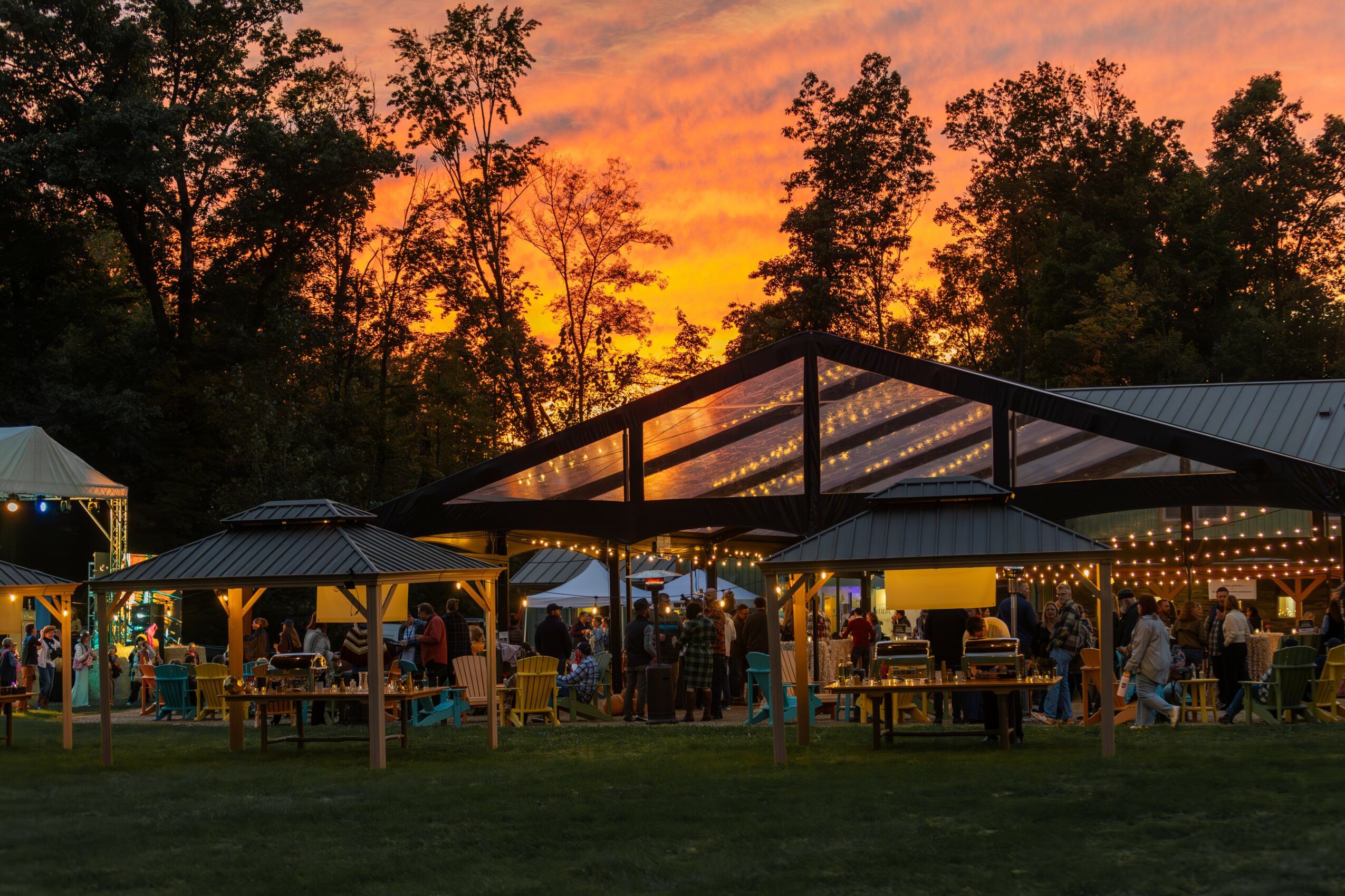 Outdoor gathering at sunset with trees, tents, and string lights, people socializing, and a colorful sky.