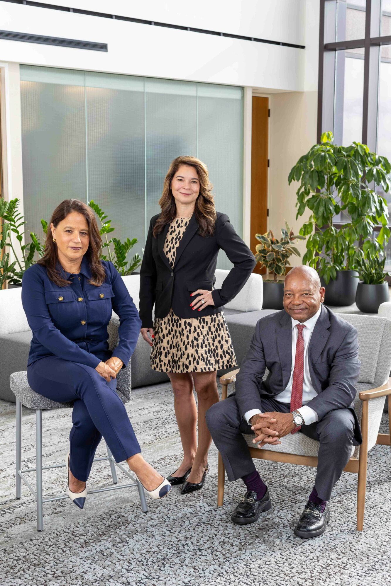 Three professionals pose in a modern office with large windows and green plants, two women and one man smiling.