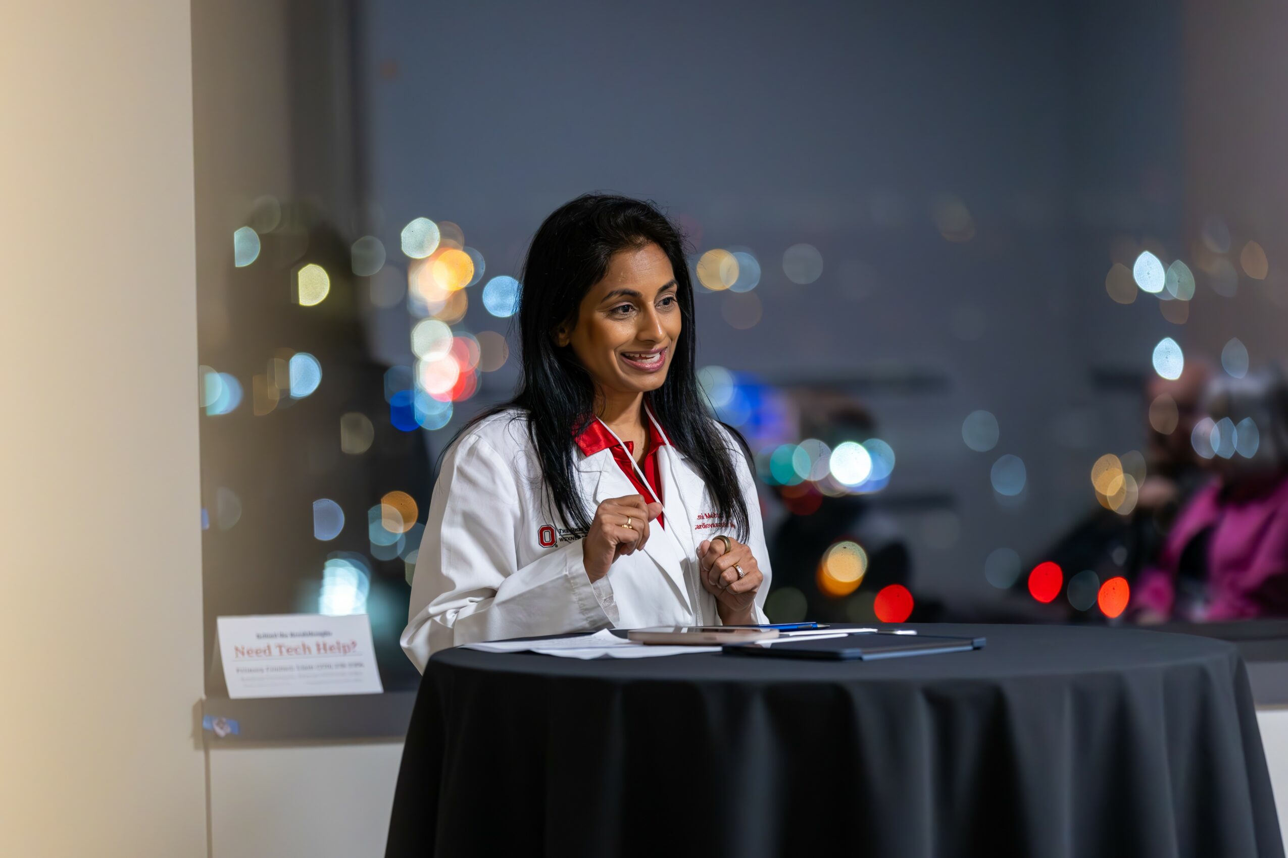 A woman in a white lab coat smiling and writing at a table with a cityscape background at night.