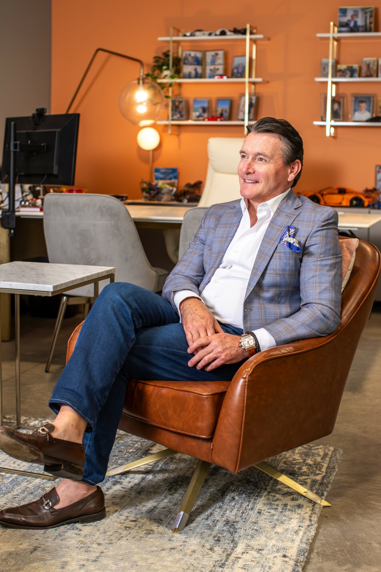 Man in a gray blazer and jeans sitting in a brown leather chair in a modern office with orange wall, shelves, and computer