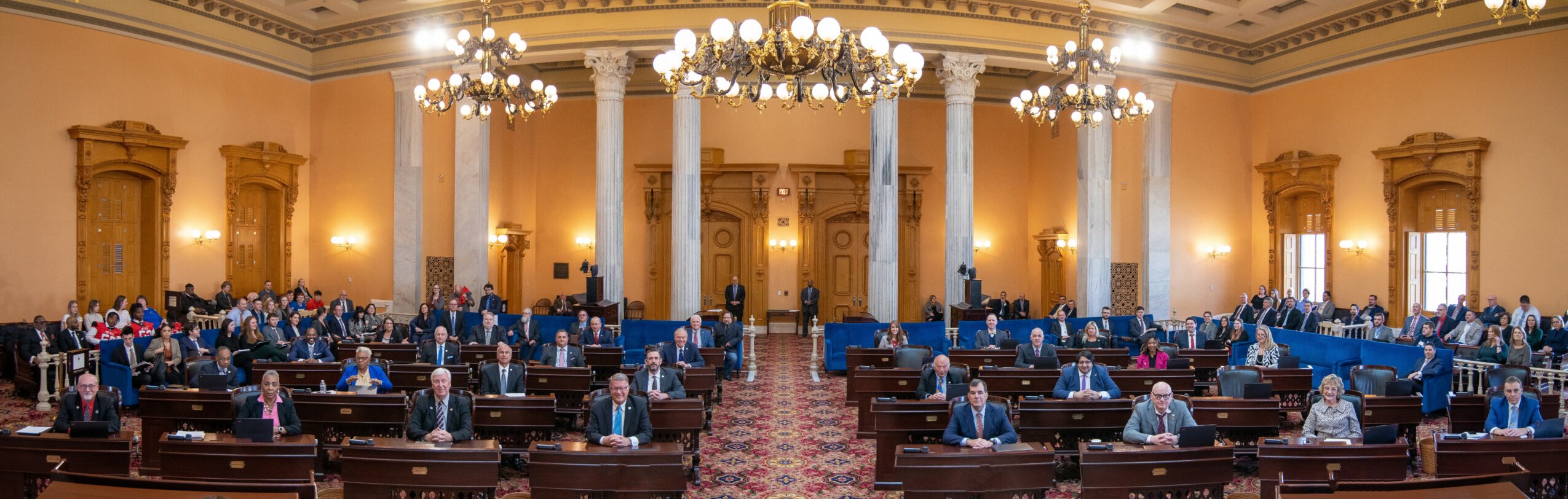 Large formal room with high ceilings, chandeliers, and rows of seats, some occupied, with ornate gold decorations and tall windows.