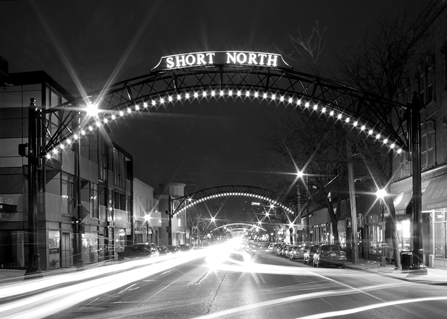 Night street scene with illuminated arches and a sign reading 'SHORT NORTH' overhead, with light trails from moving vehicles.