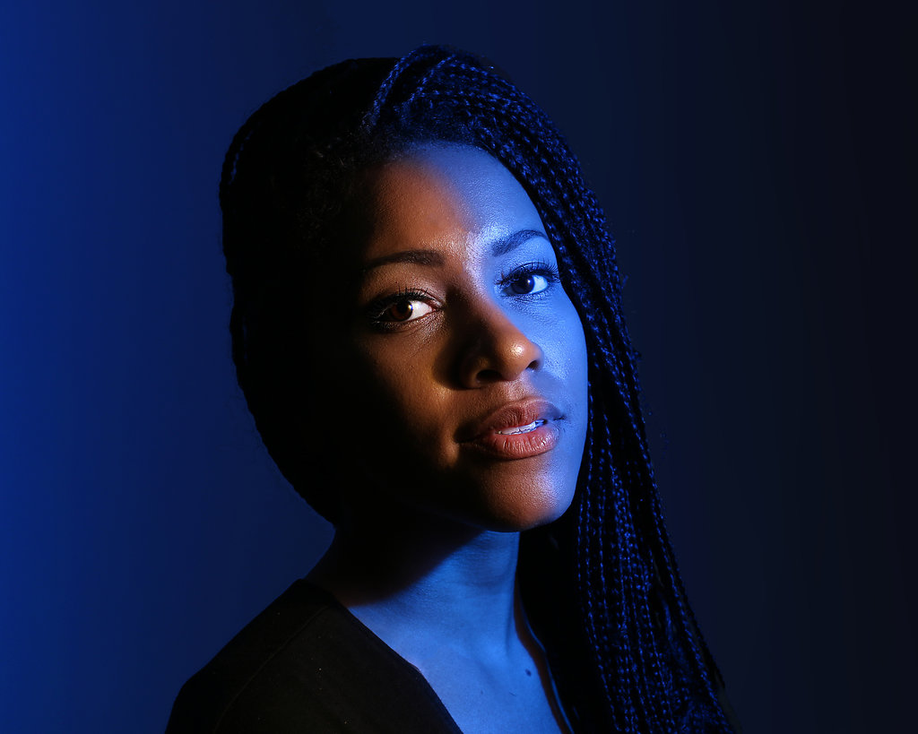 Portrait of a woman with braided hair, illuminated by blue lighting, facing slightly to the right, with a dark background.